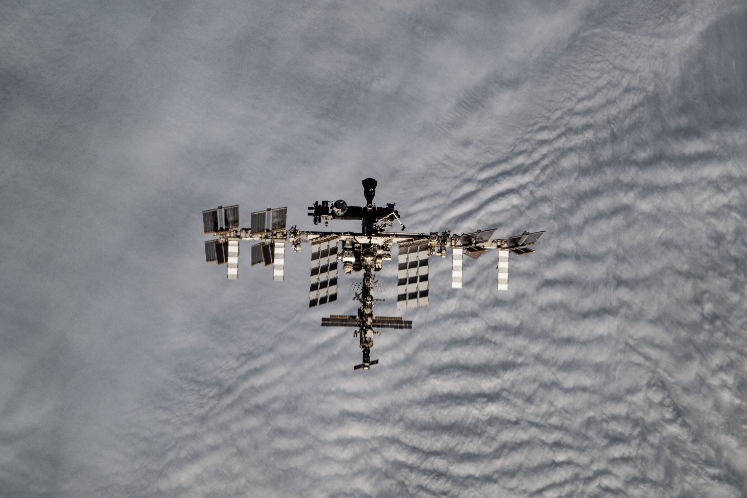 The International Space Station seen from above against rippled white clouds, its solar arrays spread along the main truss.