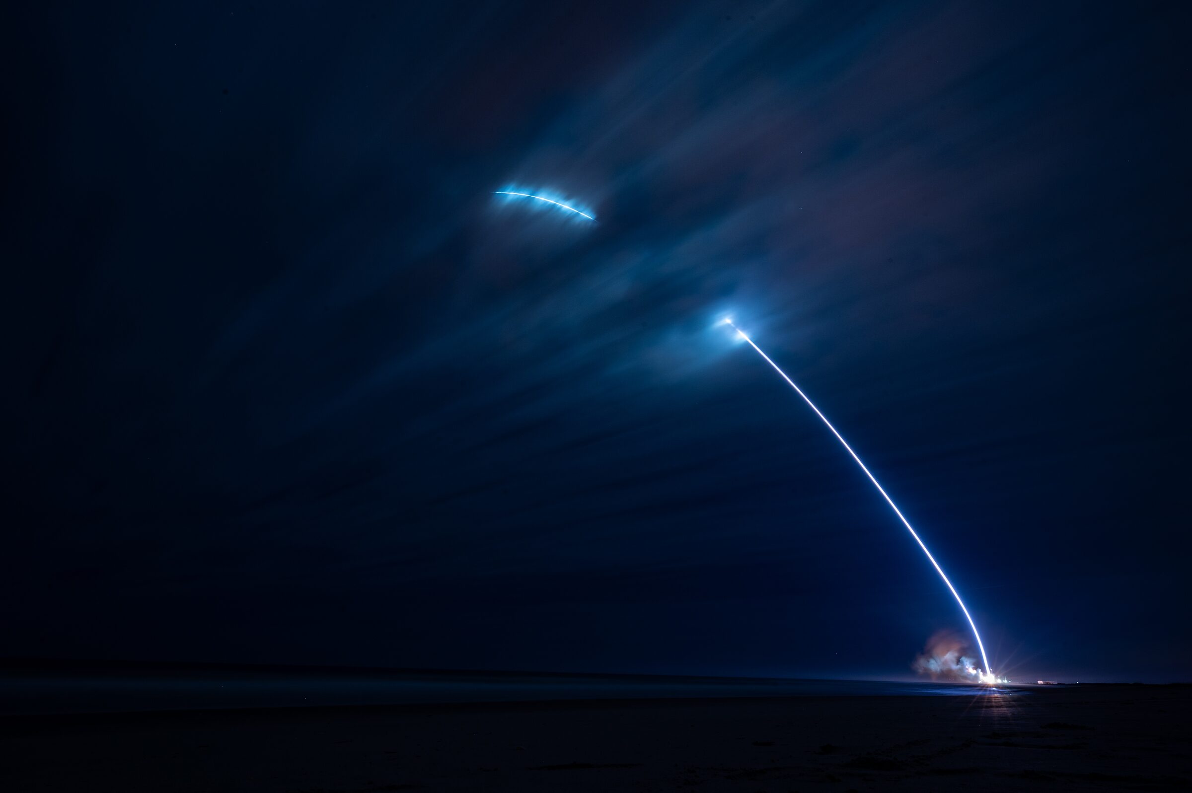 A long exposure at night: New Glenn's exhaust traces a bright arc from the launch pad at the right edge of the frame into a dark blue sky.