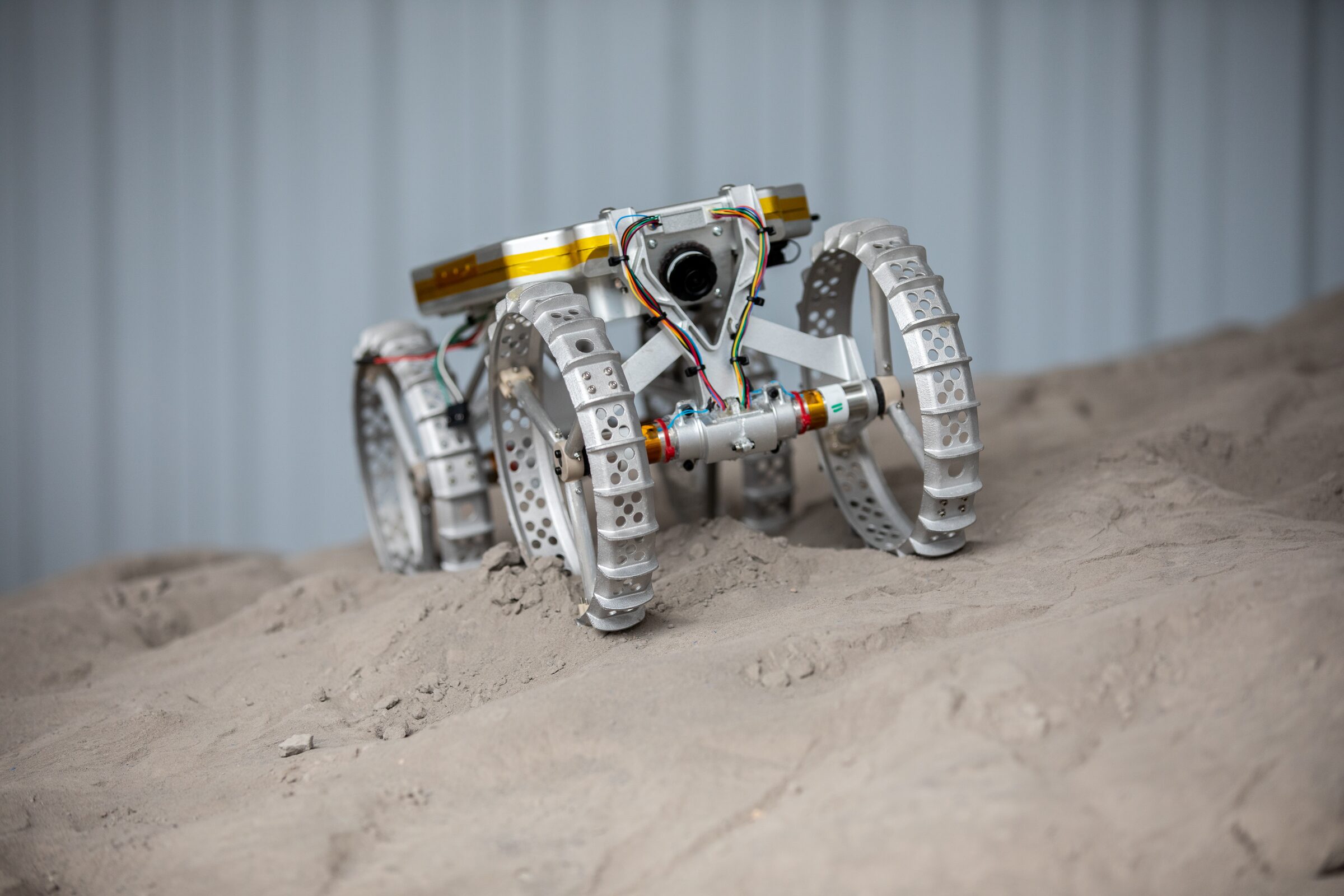 A small four-wheeled rover with perforated metal wheels and a gold-wrapped body climbs a slope of gray lunar soil simulant.